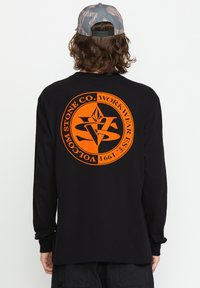 Schwarzes Langarmshirt mit einem großen orangen kreisförmigen Grafik und Text auf der Rückseite. Das Design beinhaltet geometrische Formen und ein Logo.