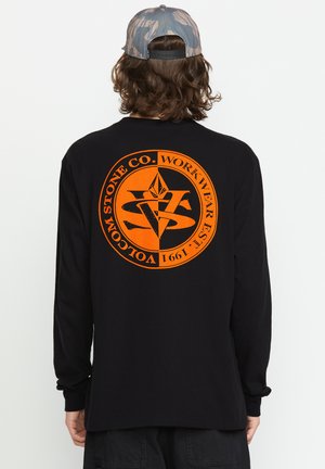 Camisa de manga larga negra con un gran gráfico circular naranja con texto en la parte posterior. El diseño incluye formas geométricas y un logotipo.