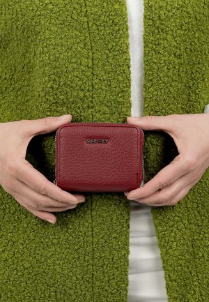 Manos sosteniendo una pequeña cartera de cuero rojo con textura y cremallera, con el logo "SURI FREY", sobre un fondo de tela verde texturizada.