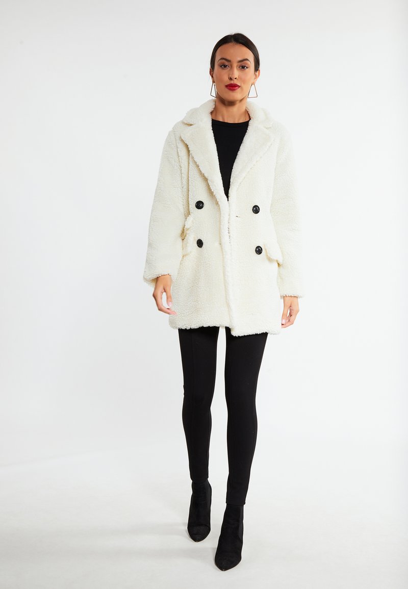 Hvid faux fur dobbeltspændt frakke med sorte knapper, med en notch-krave og sidelommer, båret over sorte leggings og anklerstøvler.