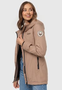 Beige, insulated jacket with a hood, featuring zippered pockets, black accents, and a circular logo patch on the sleeve.