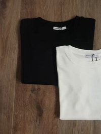 Două tricouri pliate, unul negru și unul alb, dintr-un material moale. Tricoul negru are un guler rotund, iar tricoul alb prezintă o etichetă.