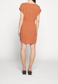 Rostfärgad klänning med vita vertikala ränder, korta ärmar, åtdragen midja och en avslappnad passform. Bärs med svarta klacksandaler med tjocka klackar.