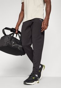 Zwarte tapered sweatpants met een elastische tailleband, gecombineerd met een licht shirt en zwarte sportschoenen. Een zwarte sporttas wordt in één hand vastgehouden.