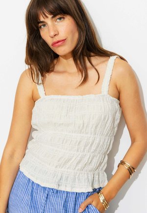 Femme aux cheveux bruns porte un haut sans manches blanc froncé, un pantalon rayé bleu et des bracelets dorés, posant sur un fond blanc.