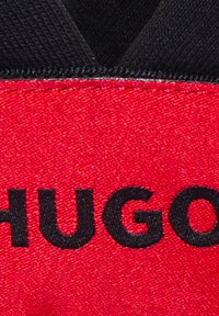 Raudona medžiaga su juodais siūlais, ant kurios didžiosiomis raidėmis parašyta "HUGO". Tekstūra atrodo lygi su šiek tiek blizgesio.