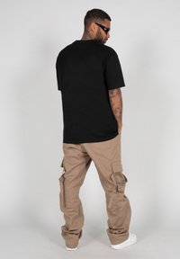 Must musta short-sleeve T-särk, millele on sobitatud tan toonis cargo püksid, millel on mitmed taskud ja lukud. Mees on vaatepunktis seljaga, kandes päikeseprille.