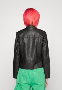 Svart skinnjacka med en kortad design, axelklaffar på axlarna och texturerad finish, kombinerad med gröna byxor för kontrast.