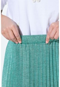 Gonna plissettata in tessuto teal chiaro con un motivo testurizzato. Vita elastica visibile, mani che afferrano la vita per la regolazione.