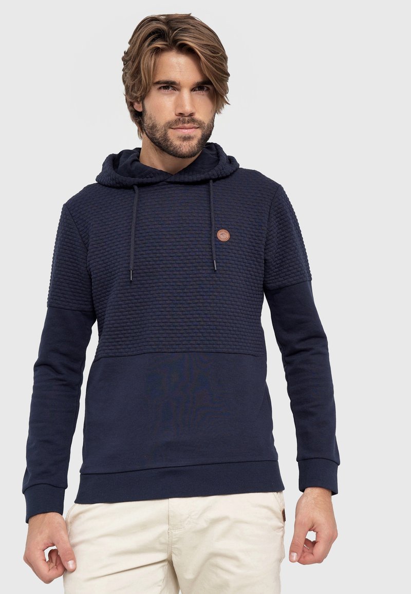 Marineblauwe hoodie met een textuur bovenpaneel en een gladde ondersectie, voorzien van een logopatch en een trekkoord voor aanpassing.
