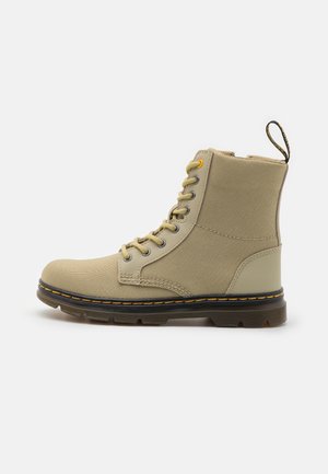 Botte beige montante à lacets avec texture en tissu, semelle noire avec surpiqûres jaunes et languette de traction à l’arrière.