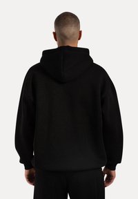 Sort hoodie lavet af blødt stof, der har en stor hætte, faldende skuldre og ribbede manchetter. Bagfra ses en afslappet pasform.