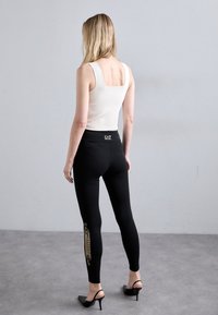 Zwarte leggings met gouden logo-details, gedragen met een witte mouwloze top. De leggings hebben een hoge tailleband en een strakke fit.