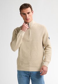 Beige, texturerad pullover med halvdragkedja, åtsittande ärmar och ribbade manschetter, kombinerad med blå jeans, bärs av en man mot en ljus bakgrund.