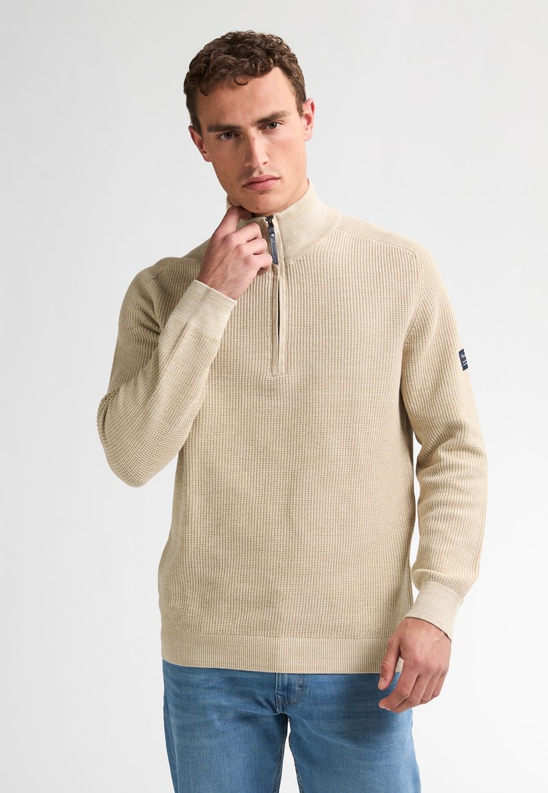 Beige, texturerad pullover med halvdragkedja, åtsittande ärmar och ribbade manschetter, kombinerad med blå jeans, bärs av en man mot en ljus bakgrund.