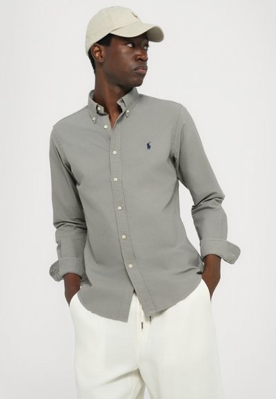 Siv srajc s prilagojenim krojem, ki ima majhen mornarsko moder logotip na prsih in svetlo bež gumbe. Model nosi bež kapo.