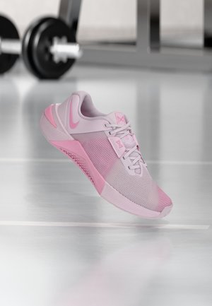 Heleroosa ja lavendli värvi Nike treeningjalanõu võrkmaterjalist pealsega ja paeltega, asetatud helkivale jõusaali põrandale, taustal ähmased raskused.