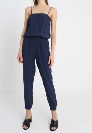 Vrouw draagt een marineblauwe mouwloze jumpsuit met elastische taille en enkels, gecombineerd met zwarte open-toe sandalen met hak.