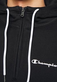 Haut à zip noir en tissu doux, avec des cordons de serrage blancs, une fermeture éclair à l'avant et un logo "Champion" en blanc.