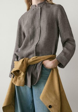 Donna che indossa una camicia grigia con bottoni, jeans blu e una giacca color sabbia annodata in vita, con una mano in tasca.