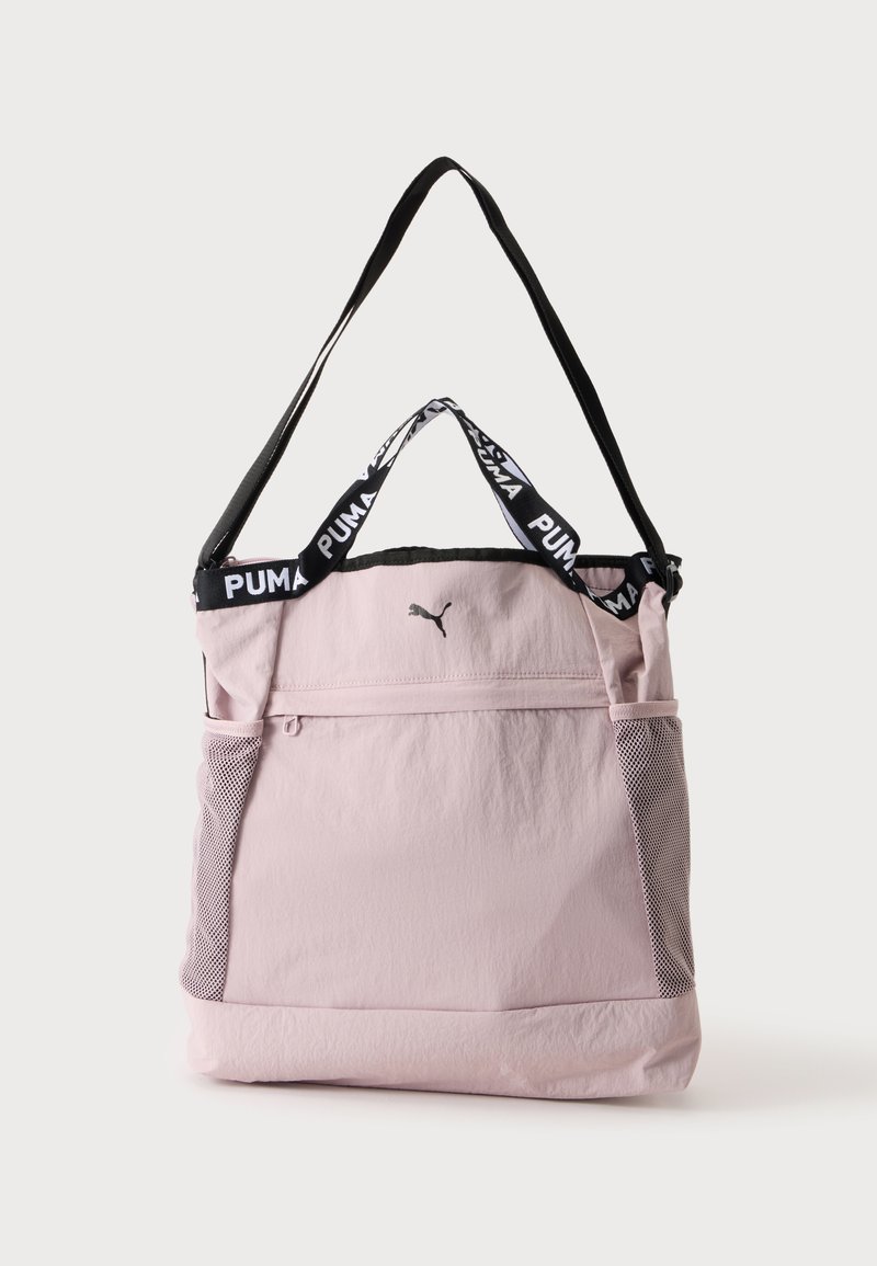 Sac fourre-tout Puma rose clair avec bandoulière noire, poignées courtes avec logo Puma, poche avant zippée et poches latérales en mesh.