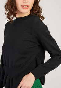 Pull en tricot noir avec un ourlet côtelé et un détail drapé sur les côtés. Comprend des manches longues et un col montant. Texture douce.