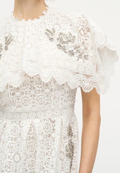 Λευκό φόρεμα από δαντέλα με κυματιστές άκρες, διακοσμημένο με ασημένιες χάντρες στους ώμους και τη μέση, που φοριέται από ένα άτομο με ακάλυπτους βραχίονες.