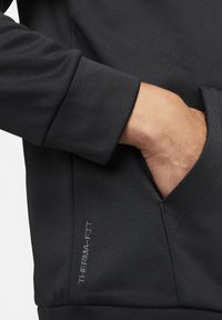 Main dans la poche d’un vêtement noir à manches longues avec le texte "THERMA-FIT" près de la couture de la poche, montrant la texture du tissu et les détails du poignet.