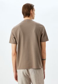 Brun t-shirt med korte ærmer i struktureret stof, rund hals og lige kant, iført med lysfarvede bukser. Set fra ryggen.