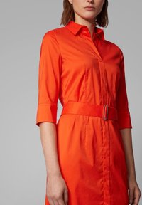 Czerwona sukienka koszulowa z zakończonym szpiczasto kołnierzykiem, rękawami o długości trzy czwarte i paskiem w talii. Gładka tekstura oraz detale szwów bocznych.