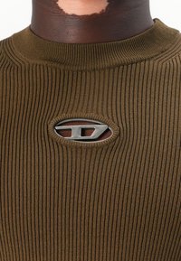 Olivegrünes, geripptes Strickshirt mit rundem Ausschnitt und einem ovalen Metallemblem in der Mitte. Die Textur ist glatt und figurbetont.