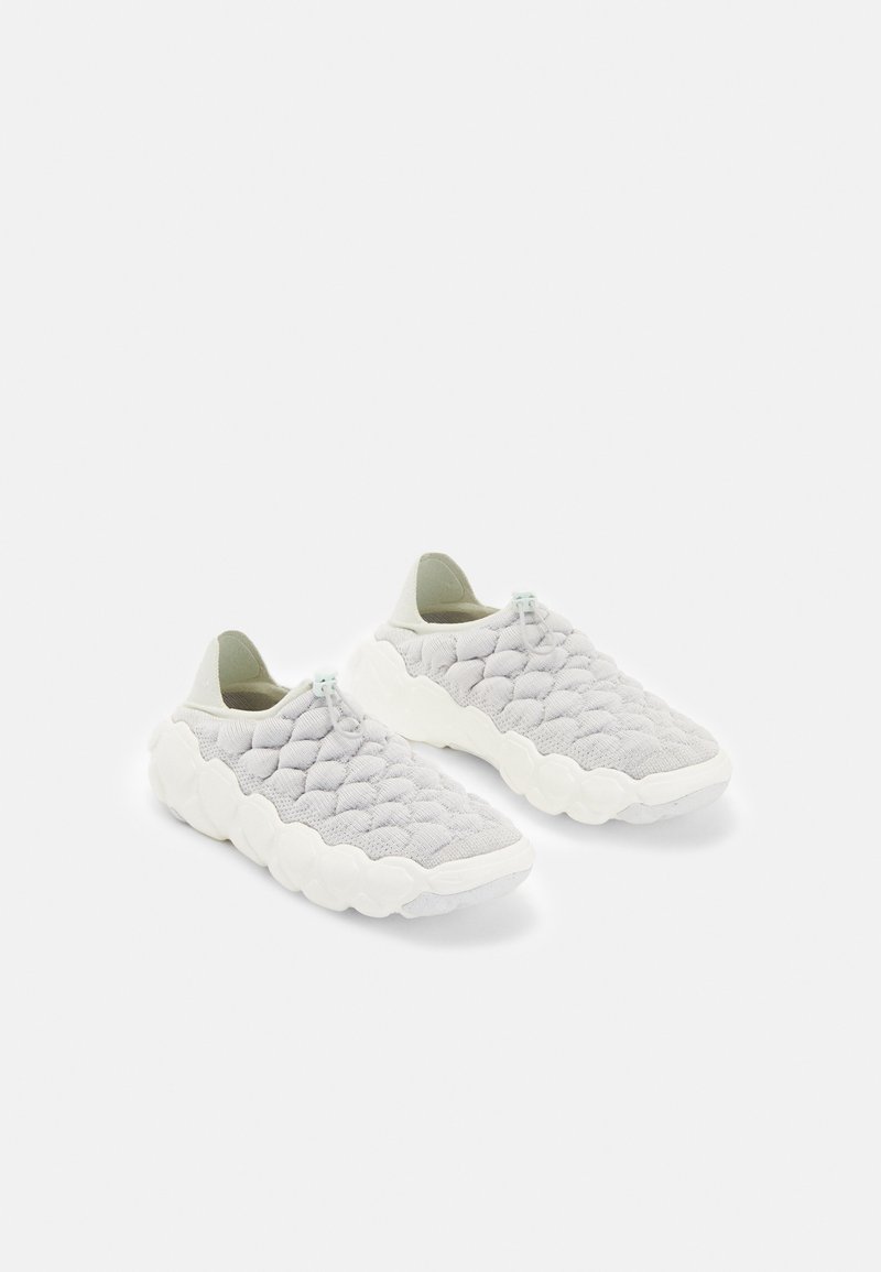 Ett par vita vadderade slip-on sneakers med tjocka strukturerade sulor och dragtabbar på en vit bakgrund.