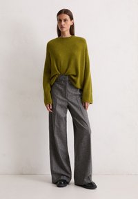 Un pull en laine vert tricoté avec des épaules tombantes, associé à un pantalon en laine gris à carreaux large et des chaussures plates noires, le tout sur un fond uni.