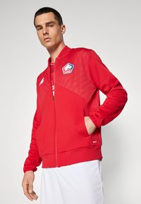 Chaqueta roja con cierre de cremallera y detalles de malla en los hombros, que presenta un logo en el lado izquierdo del pecho. Incluye puños acanalados y una textura de tejido suave.
