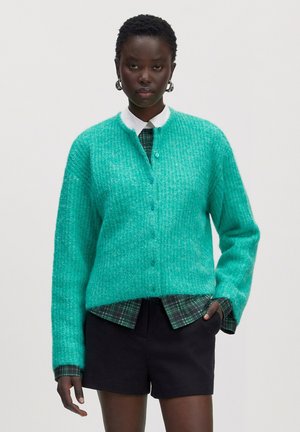Mannequin portant un cardigan boutonné bleu sarcelle sur une chemise à carreaux verte et un short noir, debout avec une main dans la poche, fond neutre.