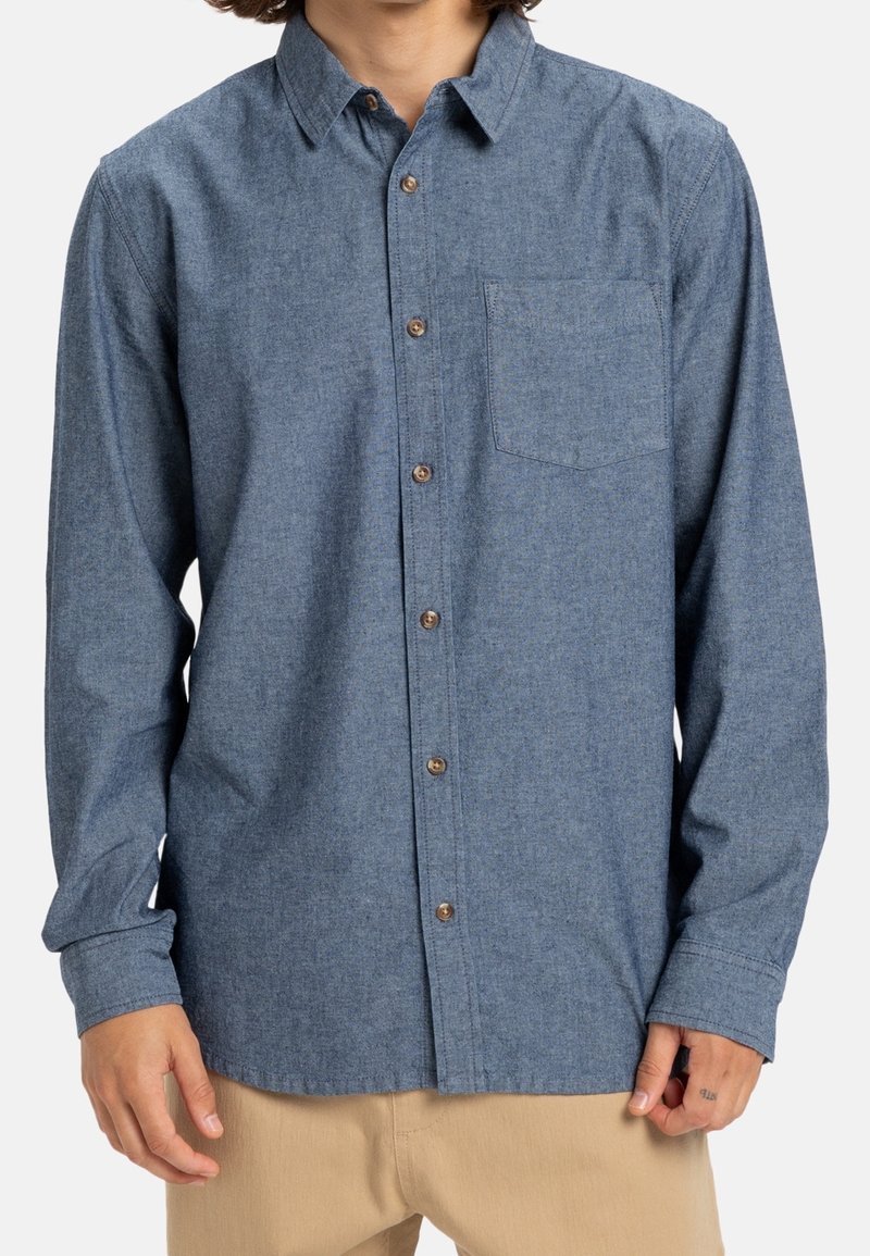 Homme portant une chemise bleue à manches longues avec une poche poitrine et un pantalon beige, debout devant un fond uni.