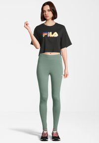 Zwarte cropped t-shirt met een meerkleurig "FILA" logo, gecombineerd met groene leggings en zwarte sneakers met roze accenten.