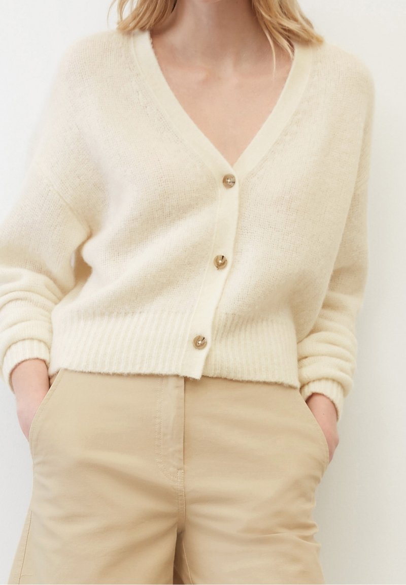 Krämfärgad stickad cardigan med V-ringning, femknappsstängning, ribbade ärmslut och nederkant. Kombineras med beiga högmidjade byxor.