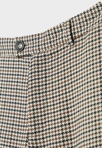 Gros plan sur un pantalon à motif pied-de-poule marron, noir et blanc, avec une taille boutonnée et des passants de ceinture.