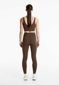 Brun sport-bh och matchande leggings, gjorda av elastiskt tyg. Leggings har rynkade detaljer vid midjan. Vita träningsskor.