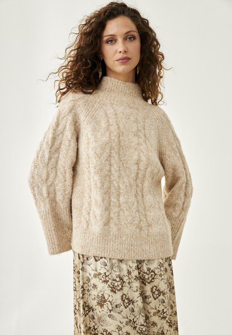 Suéter de punto beige con cuello alto; mangas anchas; tela suave y texturada; llevado sobre una falda con estampado floral en colores suaves.