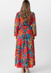 Femme avec de longs cheveux ondulés, portant une robe maxi longue à manches longues florale colorée et des bottines beige, se tenant devant un fond uni.
