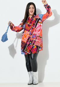 Kleurvolle, gemusterde jurk met lange mouwen, zwarte leggings en glanzende zilveren laarzen. De jurk heeft gedurfde bloemen- en geometrische ontwerpen.