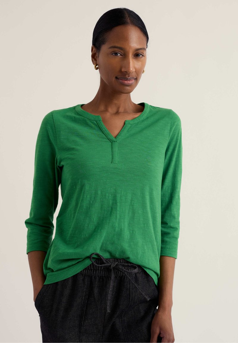 Žena s tmavými vlasmi, ktorá má na sebe zlaté kruhové náušnice, zelený top s tromi štvrťami rukávov a čierne nohavice so sťahovacou šnúrkou, stojaca na jednoduchom pozadí.