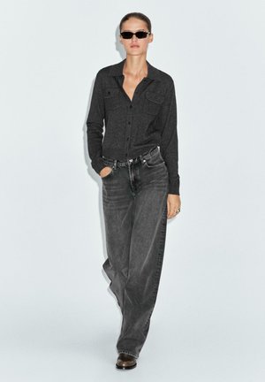 Camicia nera con bottoni e due tasche sul petto, abbinata a jeans grigi larghi. Gli accessori includono occhiali da sole neri sottili e anelli metallici.