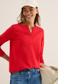 Leende kvinna med vit keps, röd långärmad skjorta och blå jeans, som håller en beige väska mot en enfärgad bakgrund.