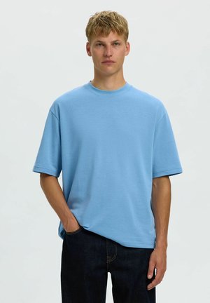 Jeune homme aux cheveux blonds courts portant un t-shirt bleu clair ample et un jean foncé, debout avec une main dans la poche sur un fond clair uni.