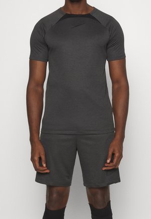 Mann, der ein dunkelgraues, kurzärmliges Sportshirt und passende kurze Hosen mit schwarzen Socken trägt und vor einem einfarbigen hellen Hintergrund steht.