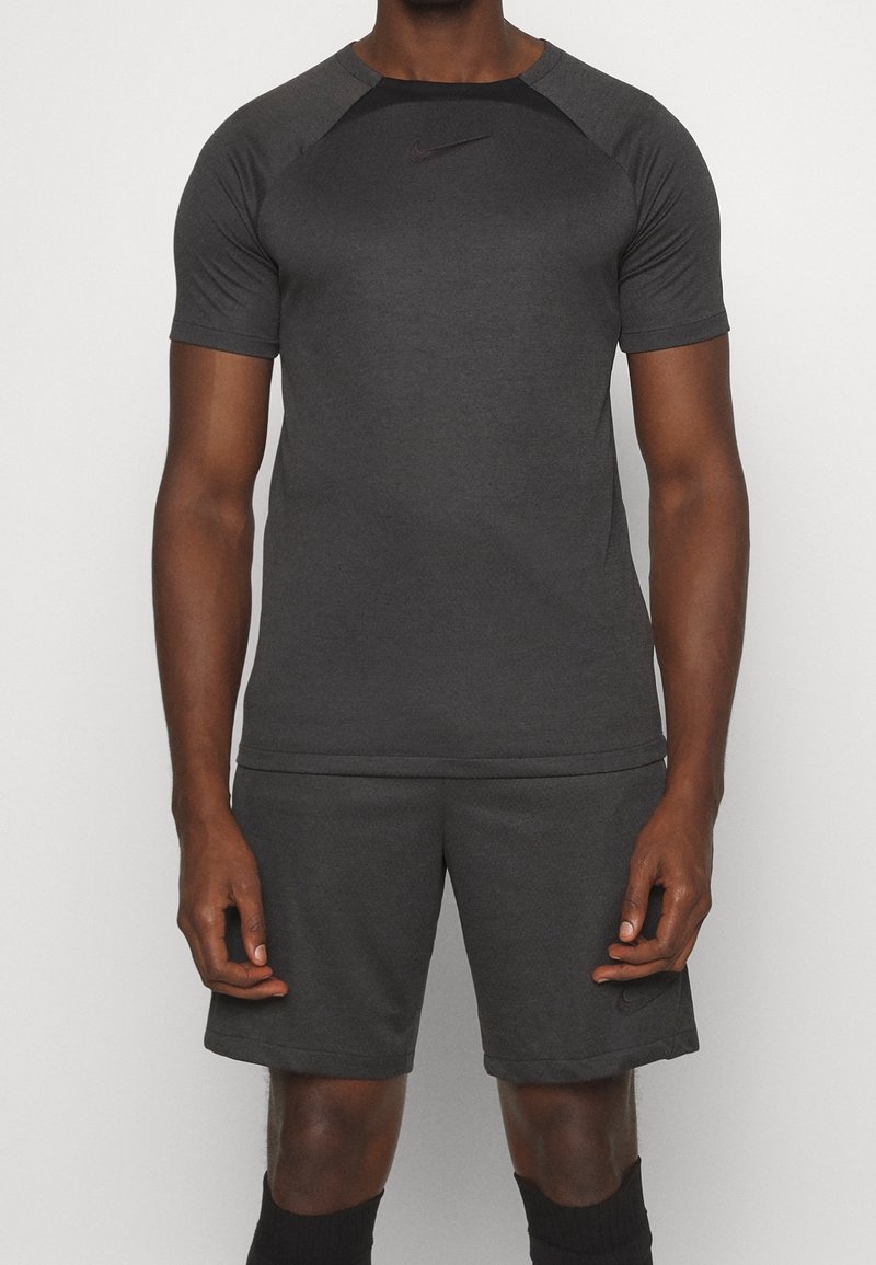 Hombre con camiseta atlética de manga corta color gris oscuro y pantalones cortos a juego, con calcetines negros, de pie contra un fondo claro y liso.