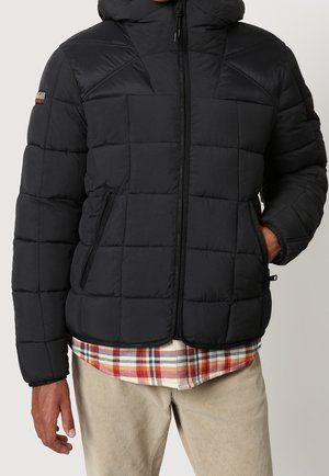 Mężczyzna ubrany w czarną pikowaną kurtkę z kapturem noszoną na czerwono-białej koszuli w kratę i beżowe spodnie sztruksowe, z ręką w kieszeni kurtki.